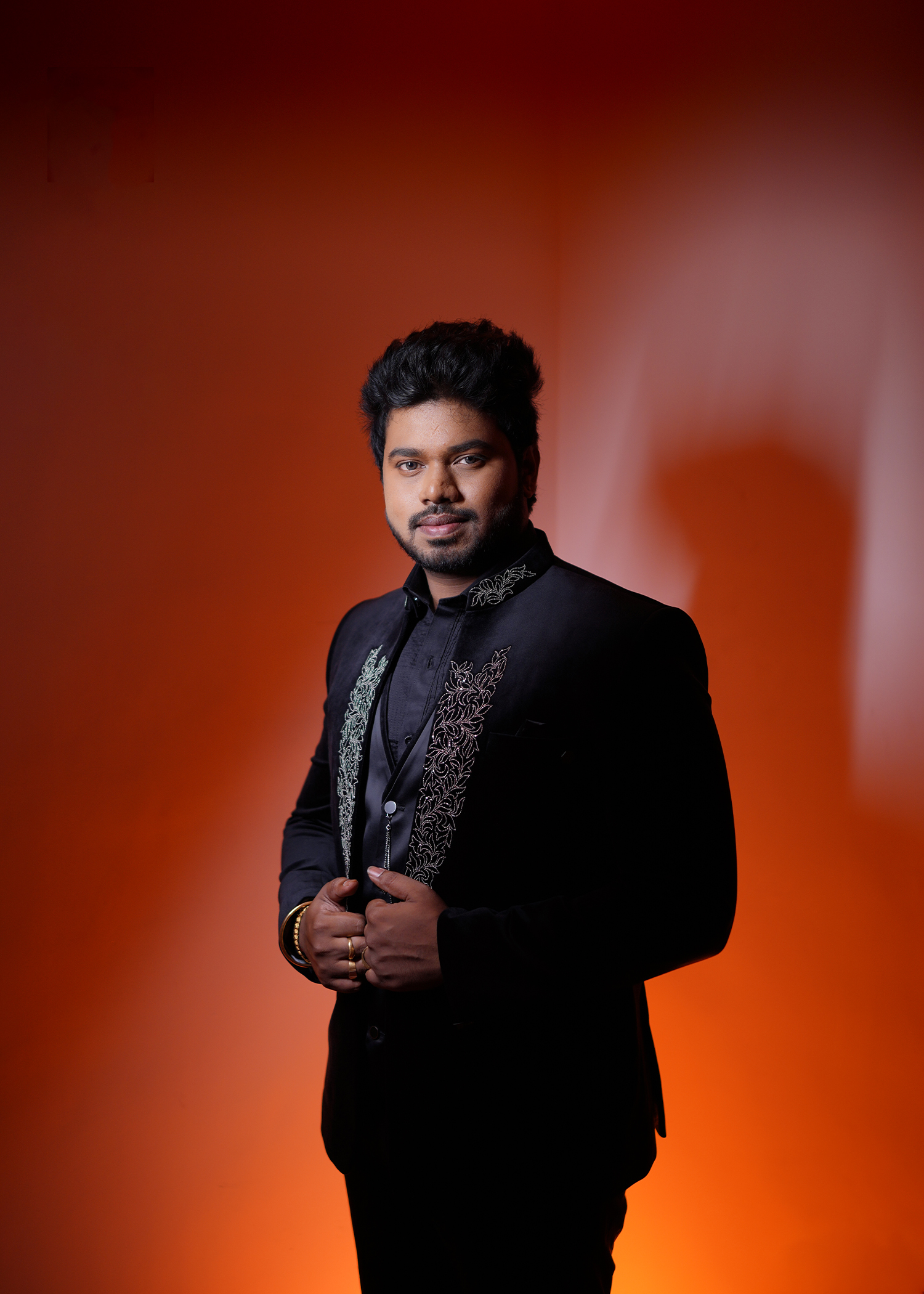 Dramatic studio portrait — subject in dark embroidered Indian suit against a warm amber-orange backdrop