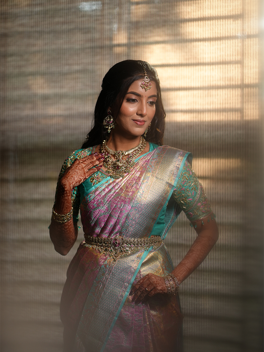 Bridal portrait — elegant bride in iridescent pink and teal silk saree with heavy gold jewellery, soft textured background